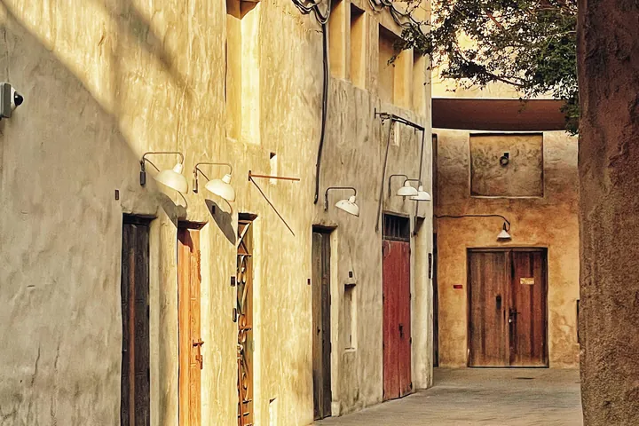 A sunlit heritage lane runs past a row of wooden doors in sand coloured walls.