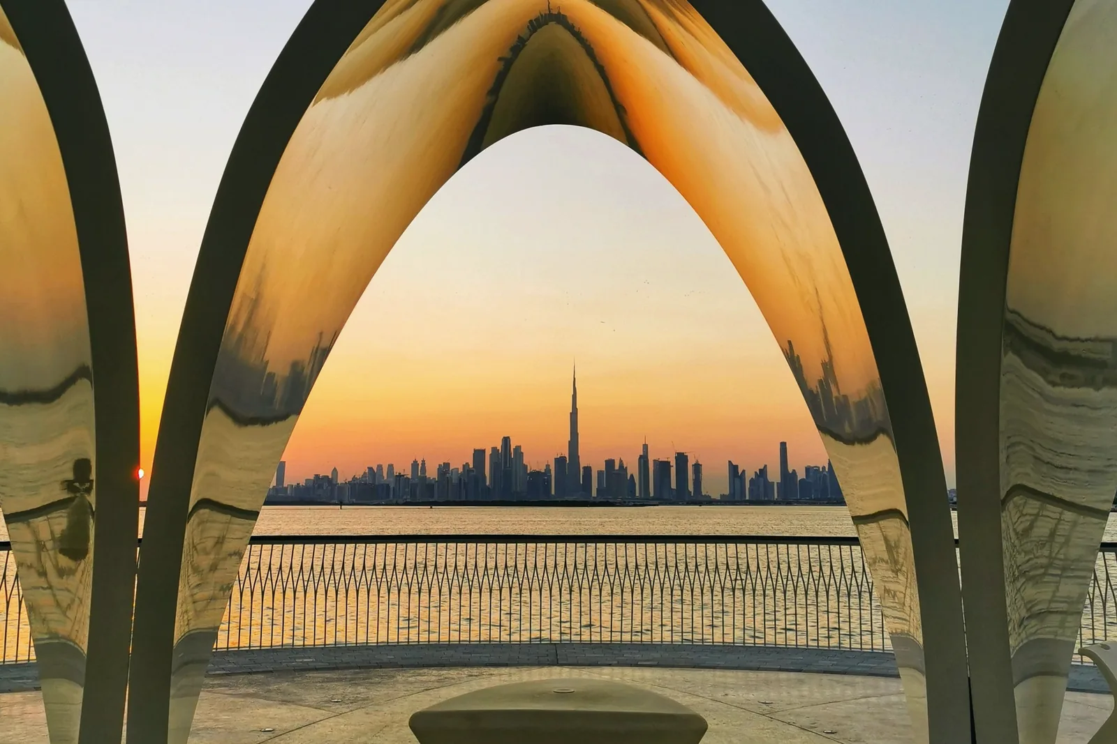 The Dubai skyline sits small on the horizon, seen through a pale arch at sunset.