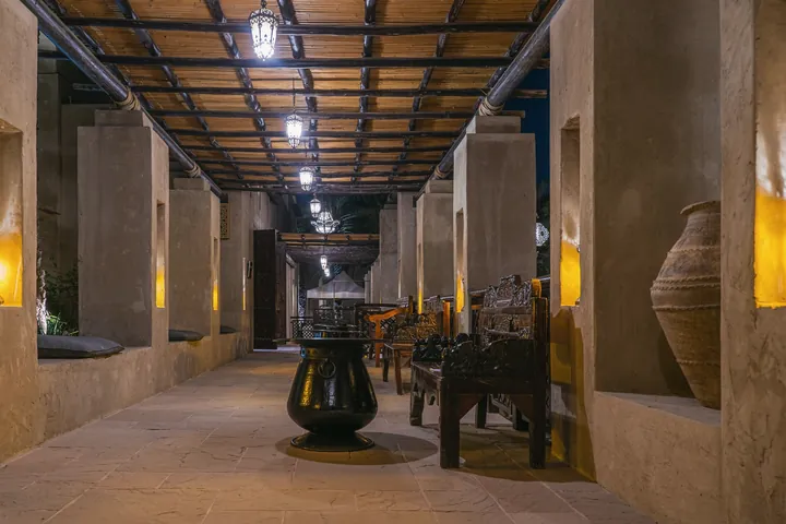 A long stone hall with carved wooden benches and a brass pot is lit by hanging lanterns.