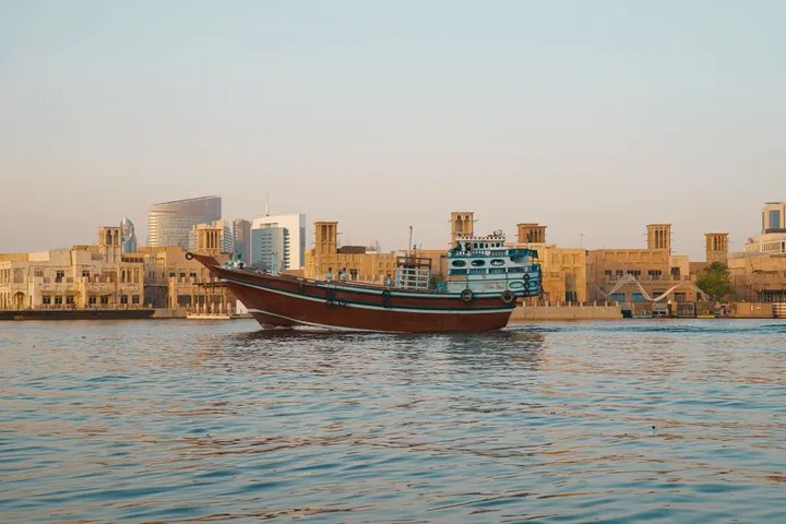 A wooden dhow passes the wind tower houses of the Creek waterfront in evening light.