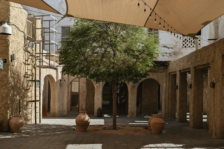 A courtyard tree stands in dappled light under sand coloured shade sails in a heritage lane.