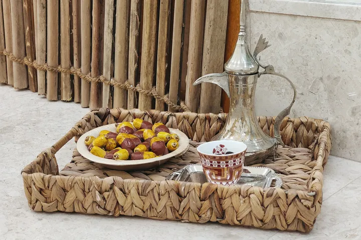 A woven tray holds a plate of dates, small cups and a brass dallah coffee pot beside a reed screen.
