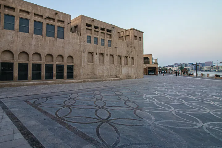 A sand coloured heritage building with a wind tower stands over a patterned stone plaza at dawn.