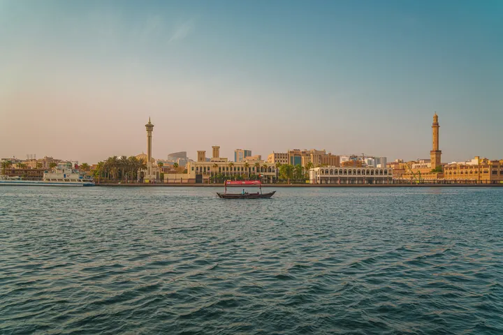 A small boat crosses the wide water of Dubai Creek towards the far bank at sunset.