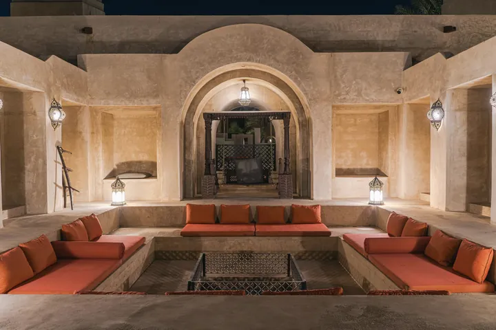 Low cushioned seating around a sunken square faces a lit arch in a stone majlis at night.