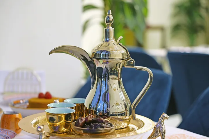 A silver dallah coffee pot, small gold cups and a dish of dates sit on a brass tray.