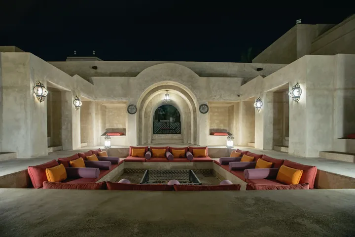 A sunken courtyard majlis at night with cushioned seating under arched walls lit by lanterns.