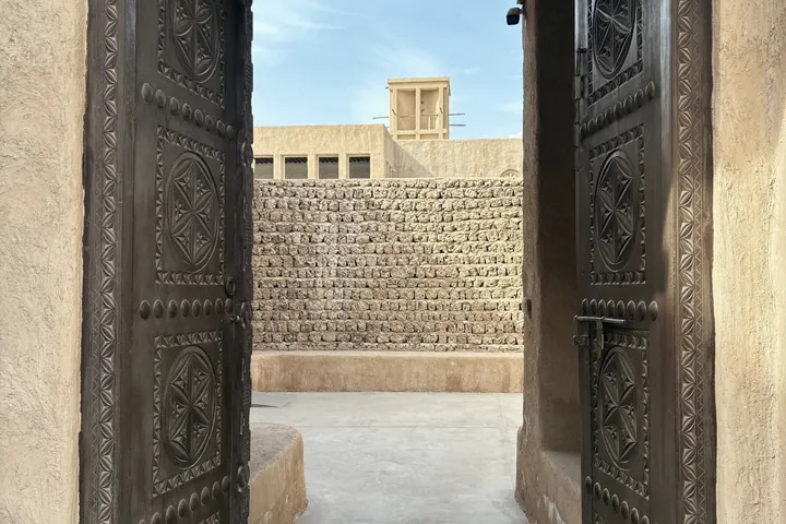 A pair of carved wooden doors stands open onto a quiet courtyard with a wind tower beyond.