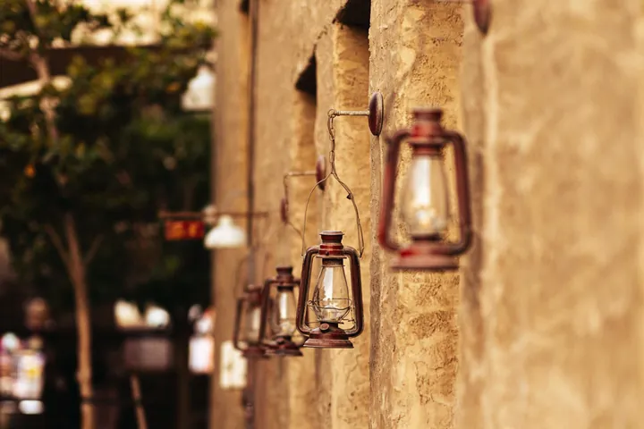 A row of old lanterns hangs along a sunlit sand coloured wall in a quiet lane.
